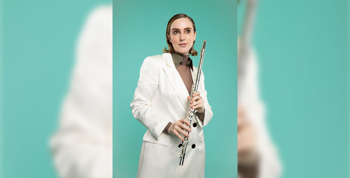 OTNES stands against a green backdrop and wears an oversized white suit, and a brown shirt with a green collar. They are holding a flute in both hands.