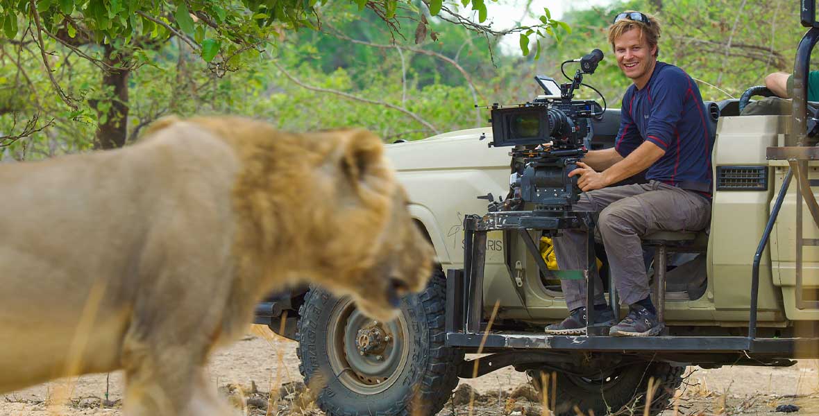 National Geographic Explorer Bertie Gregory sits in a large open-top vehicle with an attached camera dolly. He wears gray slacks and a dark blue top. He is filming a large lion in Zambia.