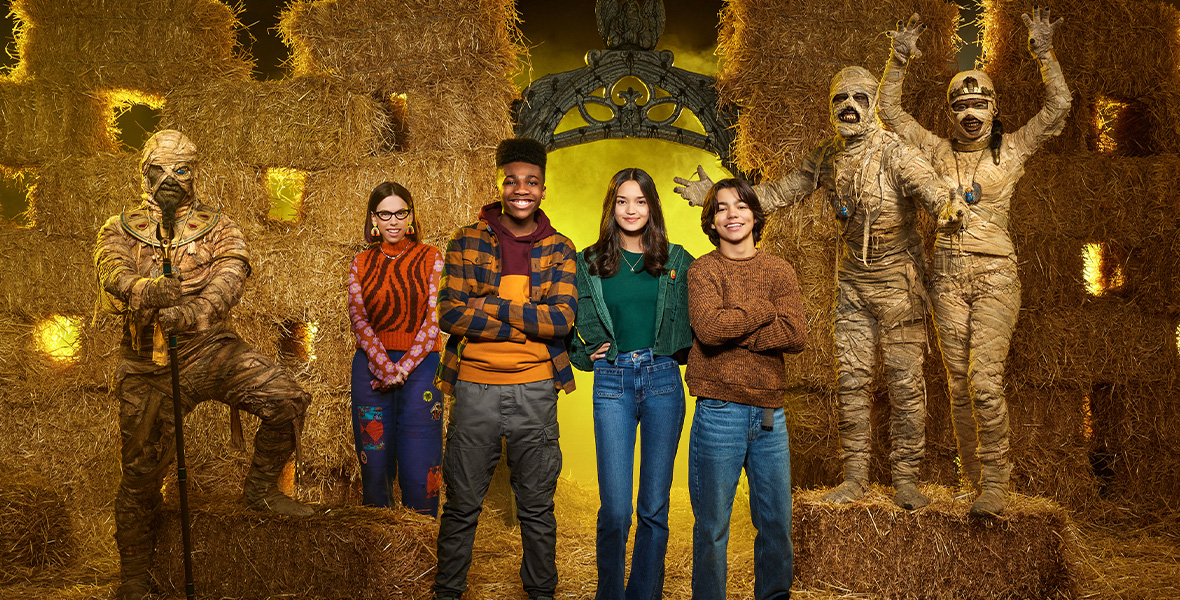 Actors Christian J. Simon, Sophia Hammons, and Malachi Barton stand center. Melanie Brook stands behind Simon. On the left, T.J. Storm portrays a mummy wrapped in gauze bandages stands with one leg on a bale of hay. On the right, Phil Wright and Rryla McIntosh portray mummies with their bodies covered in gauze bandages and stand on a bale of hay.