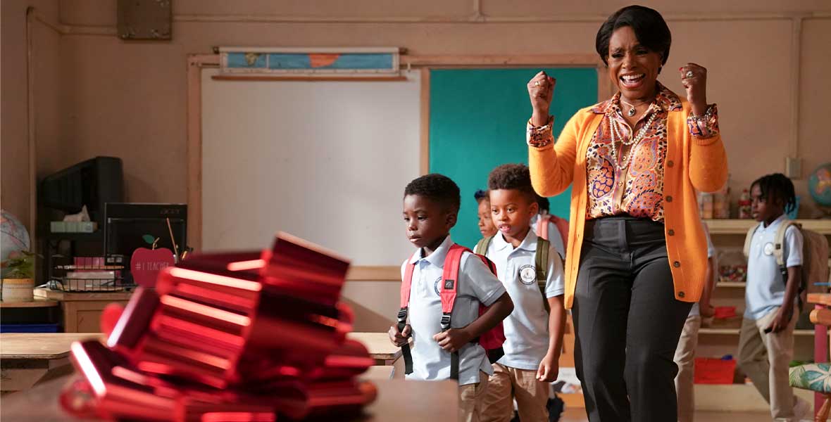 Actor Sheryl Lee Ralph pumps her arms with excitement looking at a table with new school supplies. Behind her young students are walking into the classroom with backpacks. Ralph wears an orange sweater and blouse with dark slacks.