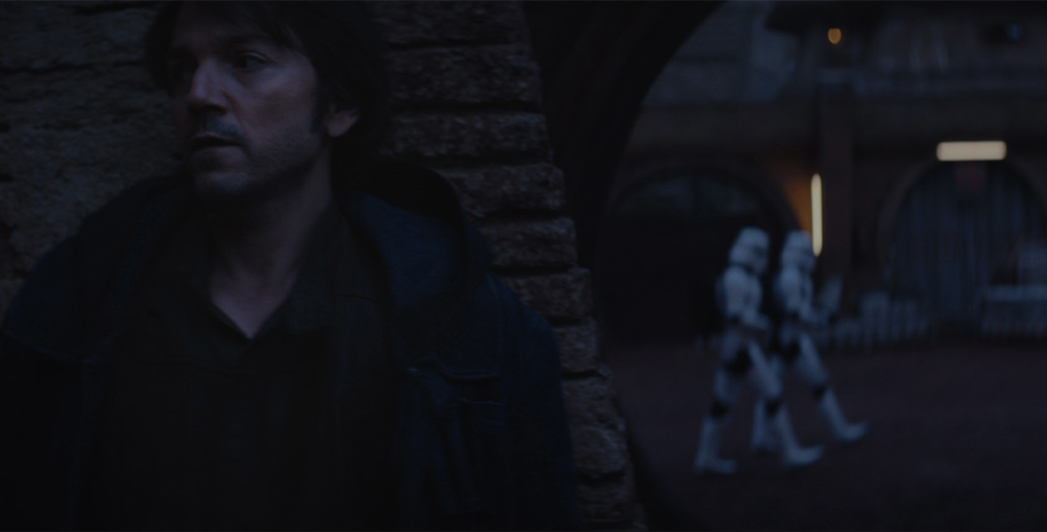 Actor Diego Luna hides behind a wall. In the distance, two stormtroopers in white armor are seen walking.