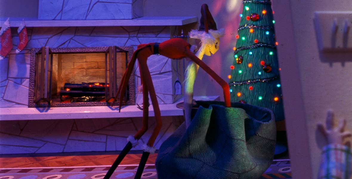 In a still from Tim Burton’s The Nightmare Before Christmas, Jack Skellington wears a Santa Claus suit and stands in front of a Christmas tree. He is bent over and reaching into a toy bag as the Christmas tree lights illuminate his face and white beard. Behind him, a fire crackles, and two stockings are hung by the chimney.