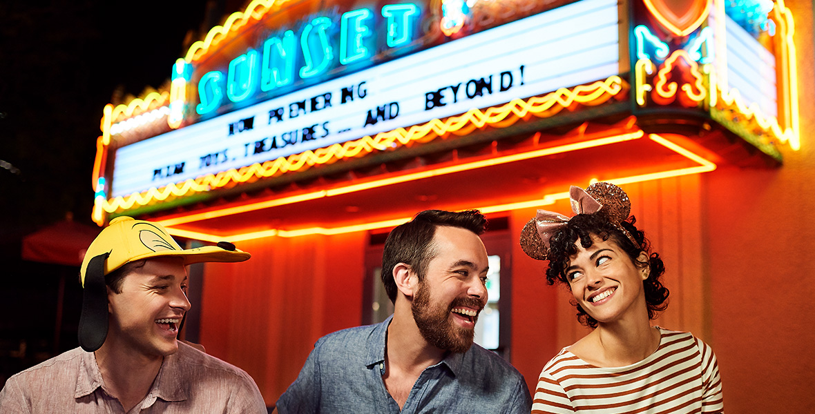 In a promotional image for Walt Disney World’s After Hours events returning in January 2023, three people are seen sitting in front of the Sunset Showcase marquee at Disney’s Hollywood Studios, at night. The man on the left is wearing a tan button-up shirt and a Pluto hat; the man in the middle is wearing a chambray shirt; and the woman on the right is wearing a cream and red striped shirt and rose gold Minnie Mouse ears, while holding a Mickey-shaped ice cream treat.