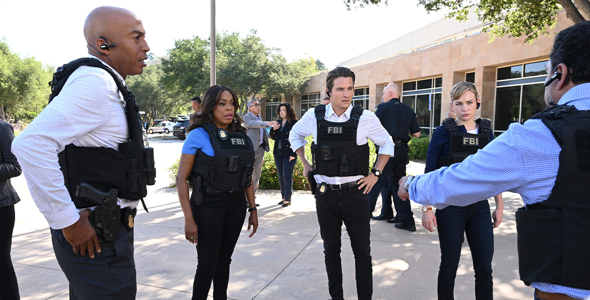 Actors James Lesure, Niecy Nash-Betts, Kevin Zegers, and Britt Robertson stand side by side outside of a brick building with large glass windows. Each wear dark pants with bulletproof vests reading “FBI”