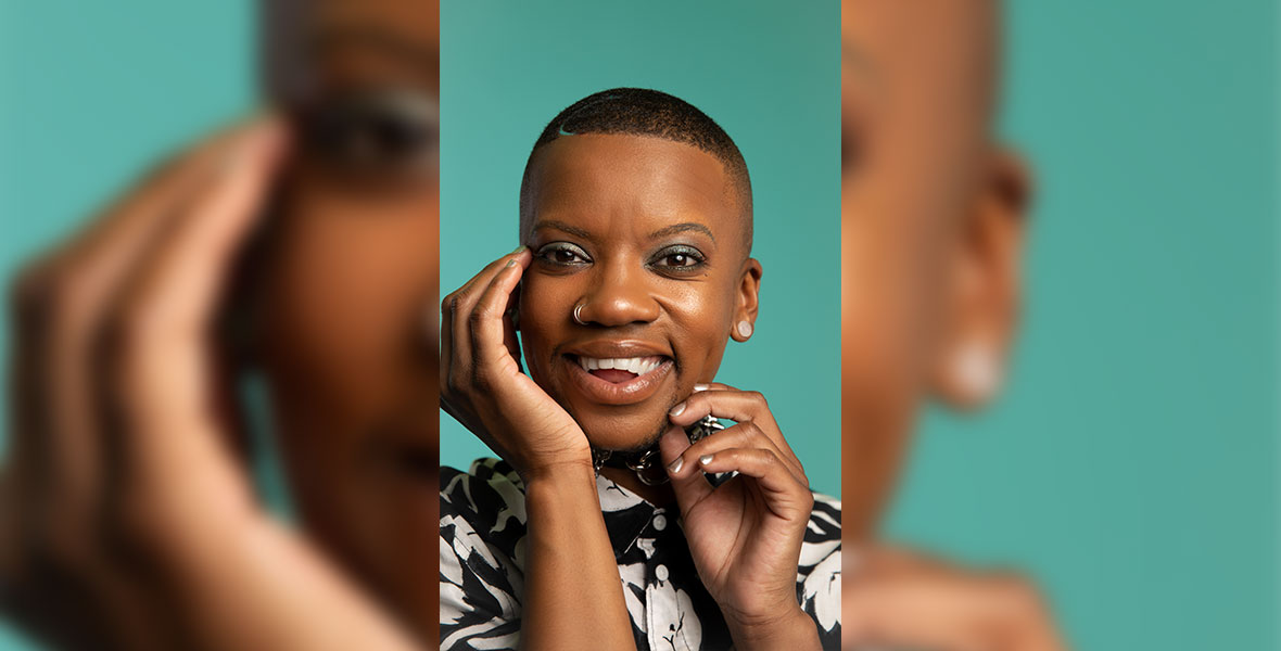 Dani Cassette poses against a green backdrop for a close-up portrait. They are smiling with their mouth open and wearing a black and white floral print shirt and two silver nose rings. They are framing their face with their hands.