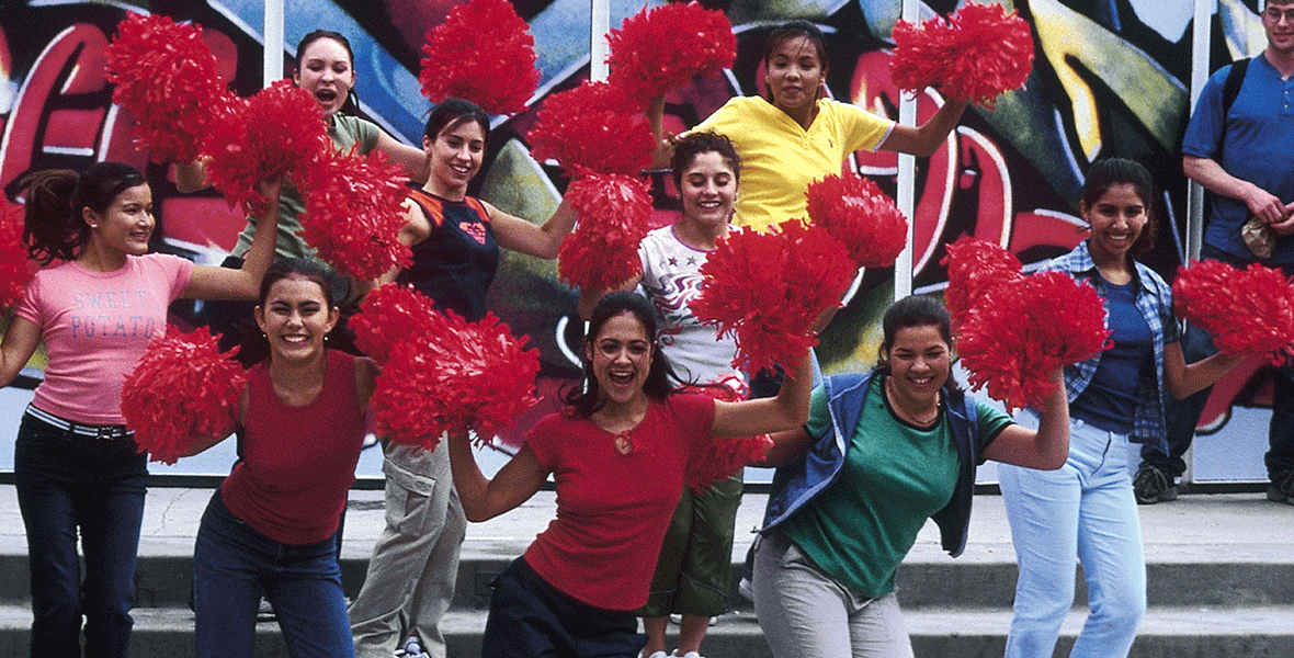 The cast is standing on concrete steps holding red pom poms in their hands with their arms next to their heads.