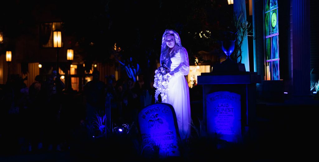 The Bride from Haunted Mansion standing on the Haunted Mansion parade float. The Bride, highlighted in purple lights, is standing in front of a replica of the Haunted Mansion surrounded by headstones. The Bride is wearing a white veil and slim white gown. The Bride is also holding white flowers that are cascading down her gown.