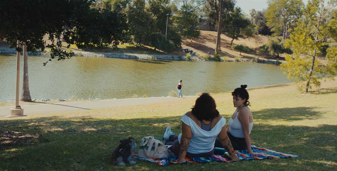 Doris Munoz and Jacks Haupt sit on a large blanket sprawled on green grass. They are leaning toward each other and face a still lake framed by green, leafy trees. Munoz and Haupt wear light-colored clothing. 