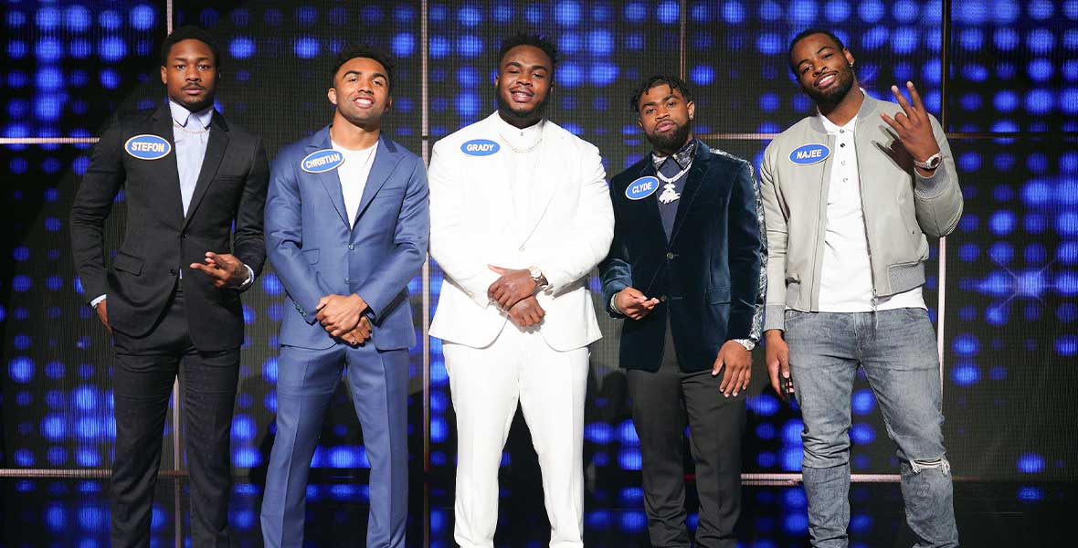 NFL football players Stefon Diggs Christian Kirk, Grady Jarrett, Clyde Edwards-Helaire, and Najee Harris stand side-by-side and in front of a large screen with alternating blue lights. All men are wearing suits and sneakers.