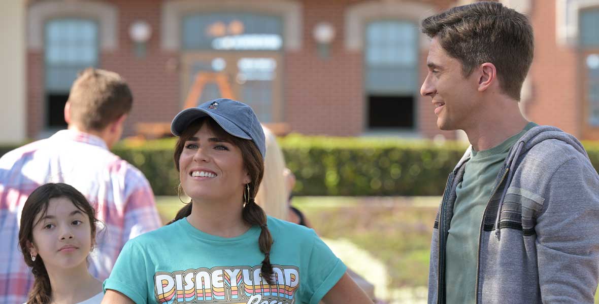 Actor Karla Souza wears a teal shirt reading “Disneyland Resort” and a gray baseball hat. She smiles with her hand on her hip and the other arm leaning on a stroller. Actor Topher Grace stands to her left and looks at her. He wears a gray hoodie and T-shirt.