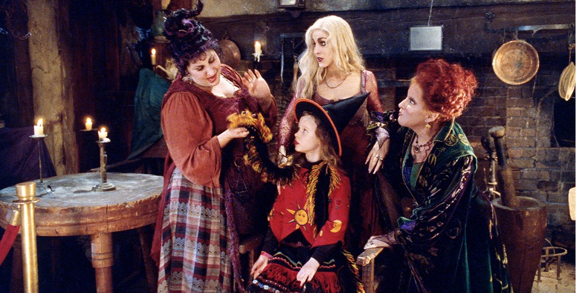 From left to right, actors Kathy Najimy, Sarah Jessica Parker, and Disney Legend Bette Midler stand around Thora Birch, who is seated in a wooden chair. Najimy wears an orange, long-sleeved shirt with a blue and orange plaid long skirt. She holds one end of Birch’s black and orange scarf in one hand. Parker looks at Najimy and wears a maroon dress with sheer, long sleeves. Midler wears an emerald green cape and gown with purple accents. Birch wears a black witch hat with a red sweater with yellow, embroidered suns.