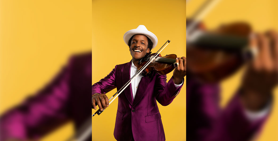 Alan Eugene Price poses against a yellow backdrop and wears a silk magenta suit and a white hat. He is playing a violin and smiling.