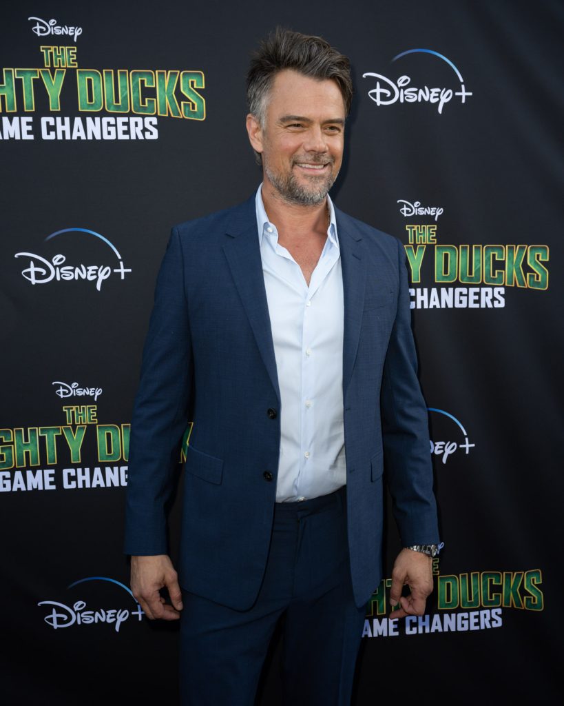Actor Josh Duhamel stands on the purple carpet at the Season 2 premiere event for The Mighty Ducks: Game Changers. He wears a blue suit with a light blue dress shirt.