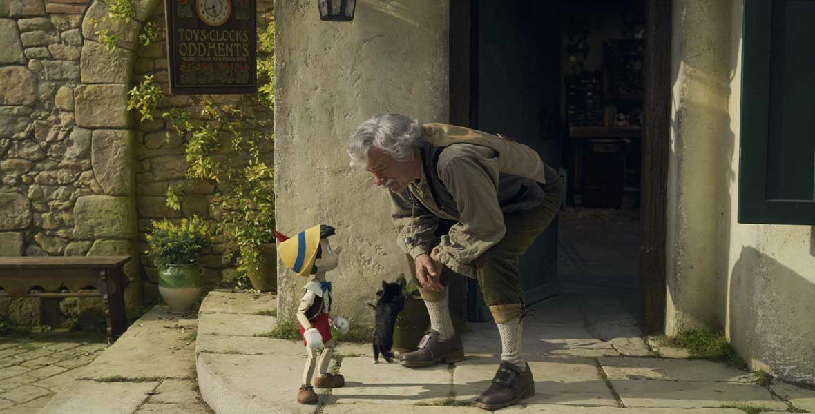 Actor Tom Hanks portraying Geppetto bends down with arms resting on his thighs to speak to Pinocchio. They stand in front of the home’s porch. A CGI depiction of Pinocchio wears a yellow hat with a single black band and a white shirt, black vest, and red shorts. Hanks wears a khaki, long-sleeve shirt and brown slacks with gray socks.