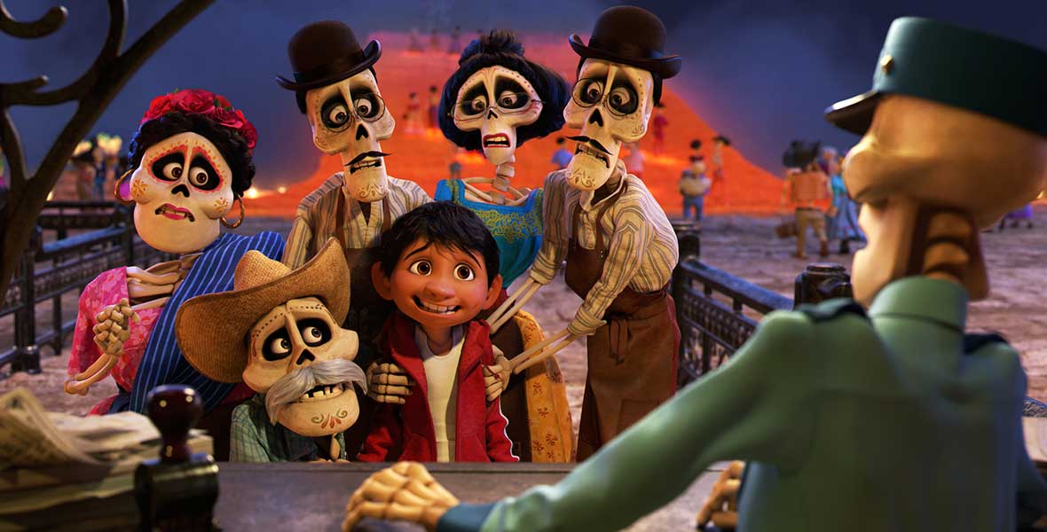 Animated boy Miguel is surrounded by a gaggle of skeletons. A large wooden desk separates the group from another skeleton, who wears a green utility hat and button-up shirt. 