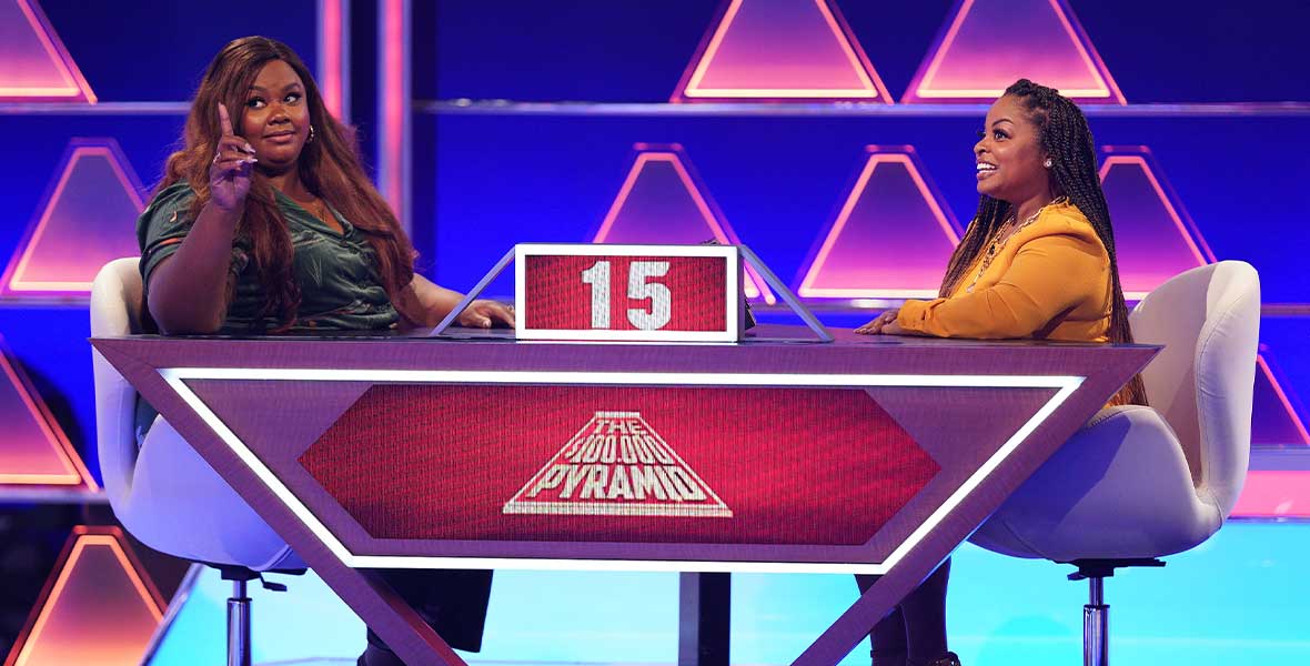 Actor Nicole Byer sits at a desk and holds up her right index finger. She wears a green, velvet ensemble. She sits across from a contestant who is looking at her. On the desk is a digital screen reading, “The $100,000 Pyramid” and a smaller screen with “15.”