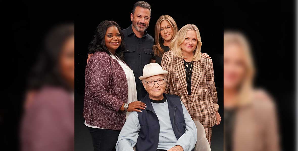 Actors Octavia Spencer, Jimmy Kimmel, Jennifer Aniston, and Amy Poehler stand behind Norman Lear who sits in a chair with his right hand on a wood table in front of him. Behind them is a black backdrop.