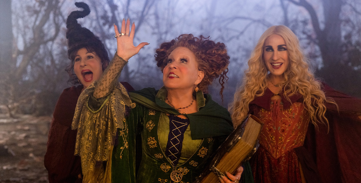 From left to right, actors Kathy Najimy, Disney Legend Bette Midler, and Sarah Jessica Parker stand in the woods. Najimy wears a dark red cloak. Midler wears a green dress and green cloak with gold accents. She holds a large, old book in her left arm. Parker wears a maroon cloak and dress with gold and green accents.