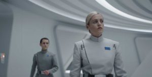 Dedra Meero stands in a white room, wearing a light-colored Imperial Officer uniform. She is frowning at something offscreen while a an officer stands behind her to the left, out of focus.