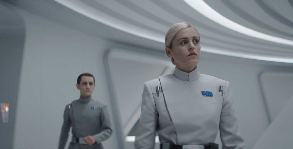 Dedra Meero stands in a white room, wearing a light-colored Imperial Officer uniform. She is frowning at something offscreen while a an officer stands behind her to the left, out of focus.
