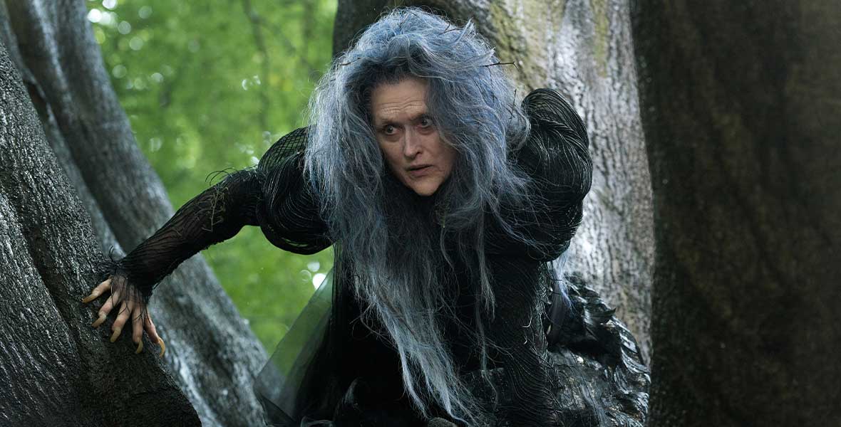 Meryl Streep, in character as The Witch, is perched in a tree. Her hair is gray and unruly, and her long nails are like talons. She is wearing an intricate black dress.