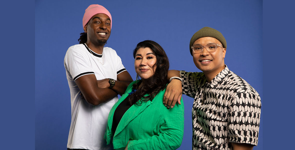 Bardo stands on the left and wears a pink beanie and a white shirt with black details. His arms are crossed and he is smiling. Caleigh Cardinal is in the center and wears a green blazer. To her left is Jeremy Passion, who wears a graphic button-up, a green beanie, and glasses. They are all smiling and posing against a blue backdrop.