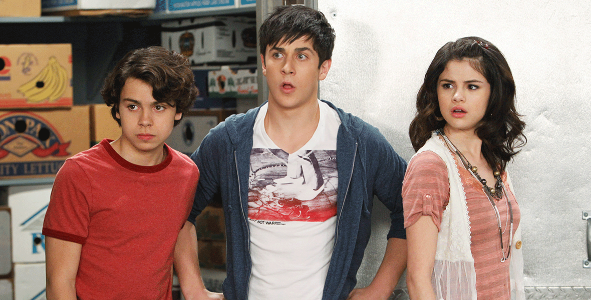 (From left to right) Jake T. Austin wears a red t-shirt. David Henrie wears a white graphic t-shirt with a blue zip-up hoodie with his hands on his hips and a surprised expression on his face. Selena Gomez stands with her right shoulder leading in a blush-colored t-shirt with a lace vest and layered necklaces.