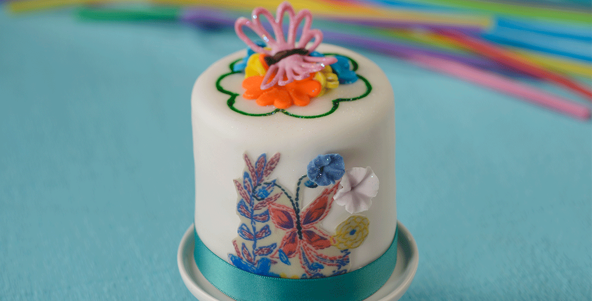 Hands holding white cake decorated with purple and red flowers and butterflies, wrapped in a teal ribbon at the bottom.
