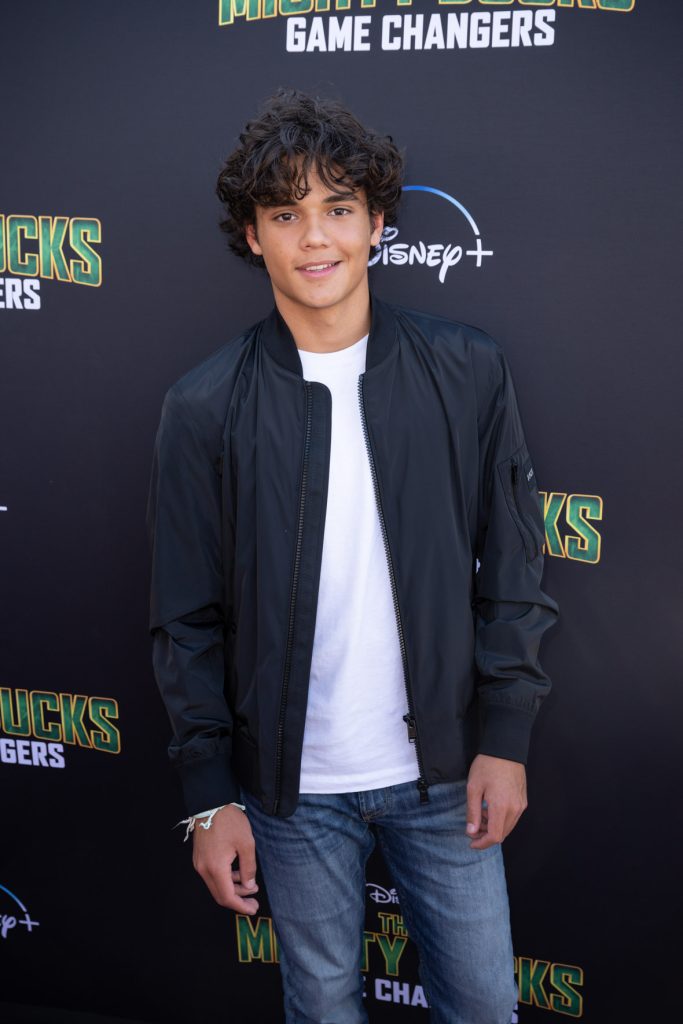 Actor Naveen Paddock stands on the purple carpet at the premiere event for the Season 2 The Mighty Ducks: Game Changers. He wears dark blue jeans with a white T-shirt and black jacket.