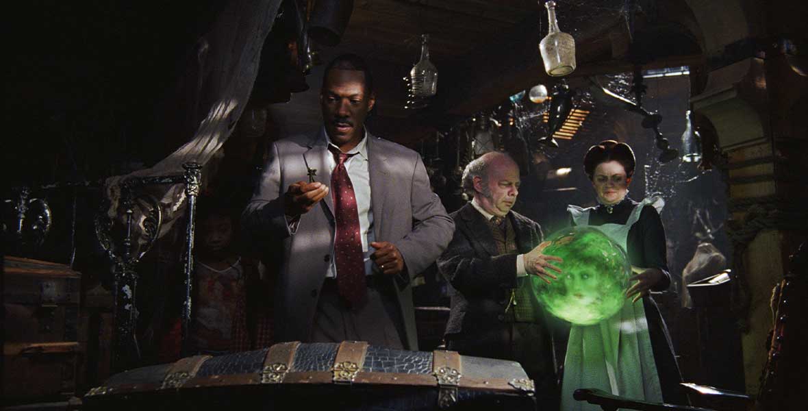 In a still from The Haunted Mansion (2003), Jim Evers, played by Eddie Murphy, inspects a key in a dimly lit room. His daughter, Megan, played by Aree Davis, stands just behind him, cloaked in shadows. Behind them are the ghost Ezra, played by Wallace Shawn, and the ghost Emma, played by Dina Spybey, who are holding a crystal ball containing the glowing green head of Madame Leota, played by Jennifer Tilly.