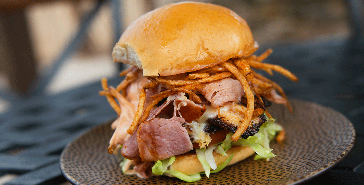 Large Burger stacked with ham, pork belly, and fried potato sticks resting on a brown plate.