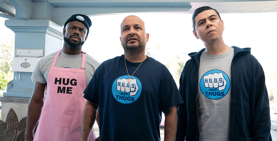 Three members of the cast of This Fool stand next to each other. The man on the left is wearing a pink apron that reads “Hug Me,” while the man in the middle (Frankie Quinones) and the man on the right (Chris Estrada) wear T-shirts reading “Hugs Not Thugs.”