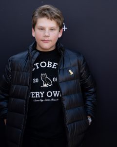 Maxwell Simkins stands on the purple carpet at the premiere event for the Season 2 The Mighty Ducks: Game Changers. He wears a black puffy jacket with a black T-shirt with white text across the front.