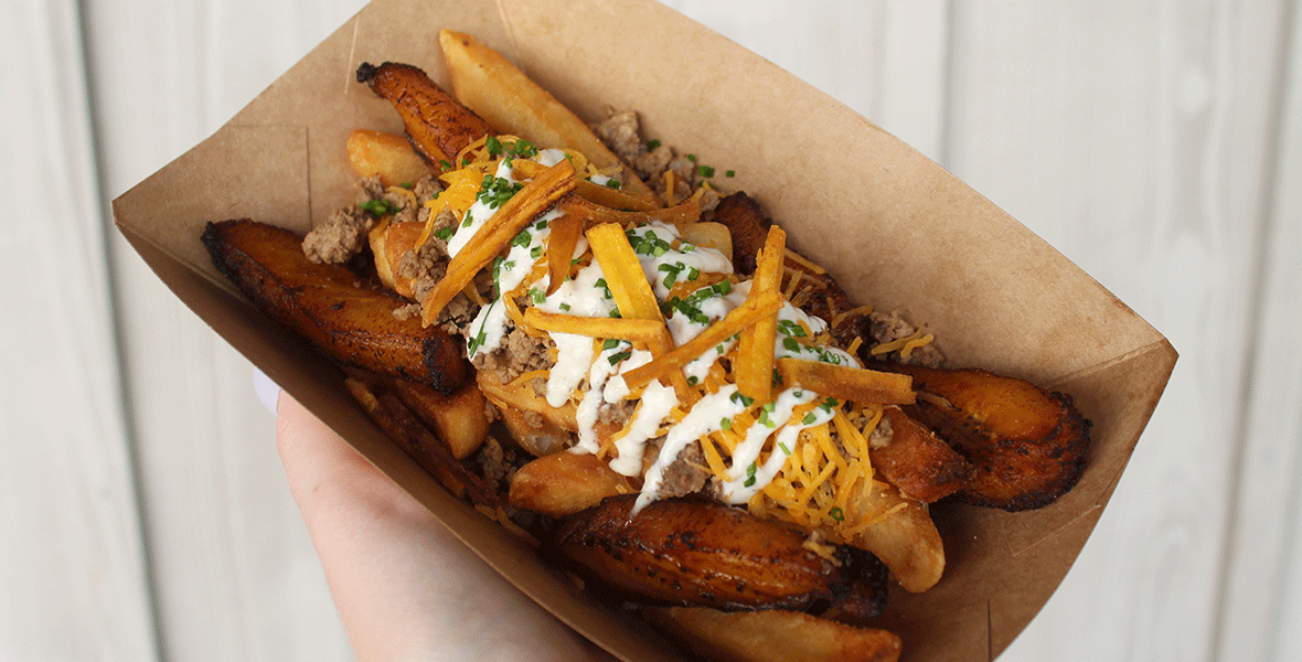 Fries topped with sofrito ground beef, fried sweet plantain, shredded cheddar cheese, garlic mayo, and chives held on a brown paper tray.
