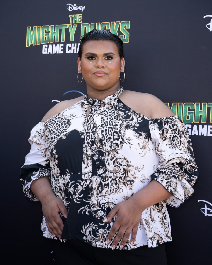 Luke Islam stands on the purple carpet at the premiere event for the Season 2 The Mighty Ducks: Game Changers. They wear a black and white patterned blouse that hangs off the shoulders.