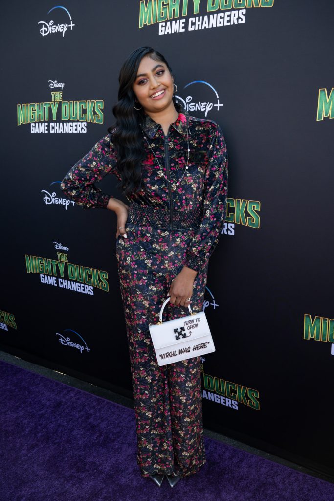 Actor Swayam Bhatia stands on the purple carpet at the premiere event for the Season 2 The Mighty Ducks: Game Changers. She wears green and pink floral-patterned jumpsuit and holds a white purse in her left hand.