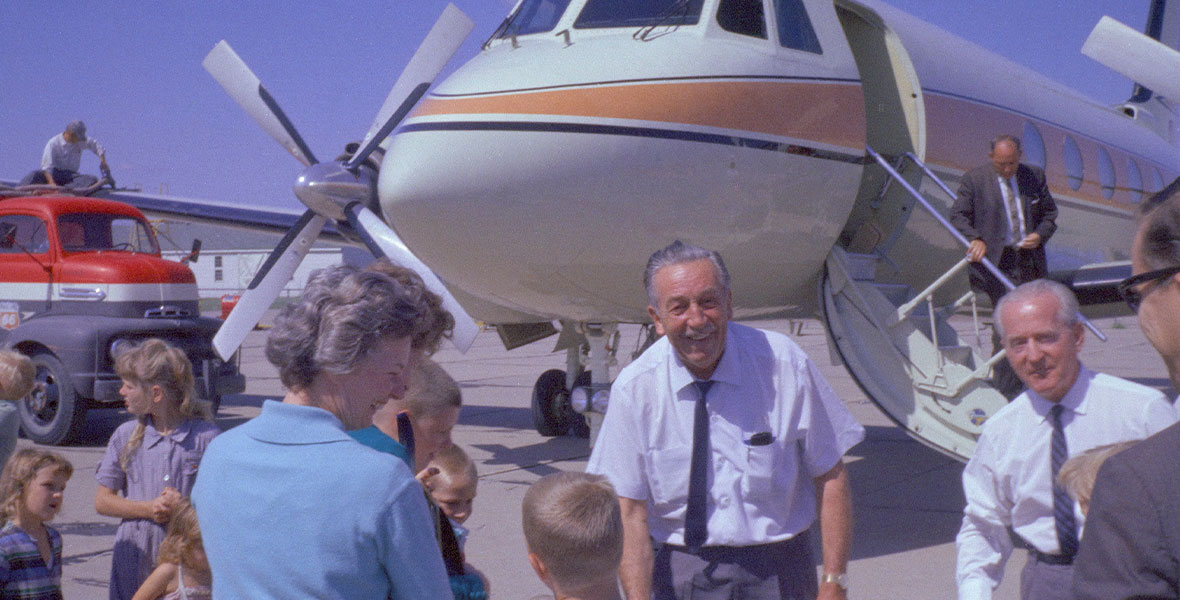 Walt Disney greets children in front of his Grumman Gulfstream I Airplane, which sits on a runaway with its door open.