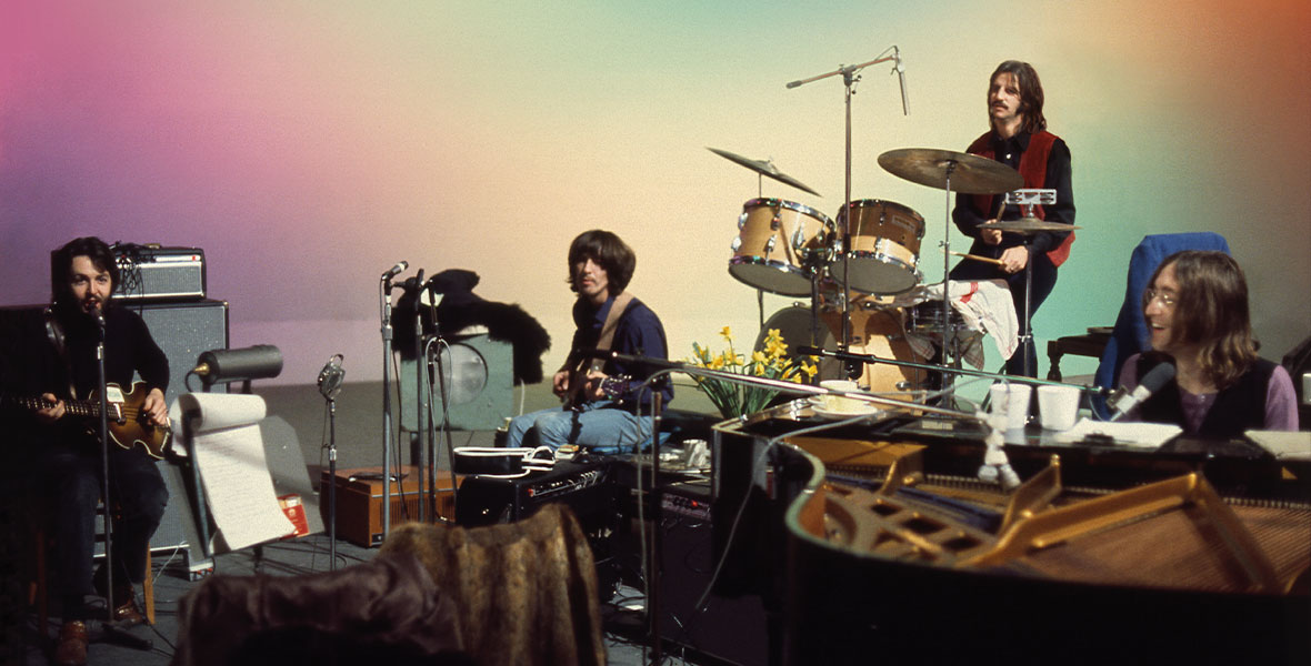 The Beatles record music in a rainbow colored room. Paul McCartney is to the left, wearing all black, and strumming his guitar. Closest to him is George Harrison, wearing jeans and a blue shirt. Behind him on drums is Ringo Starr, wearing a red vest, a black shirt, and black pants. To the right is John Lennon, who is smiling at the piano.