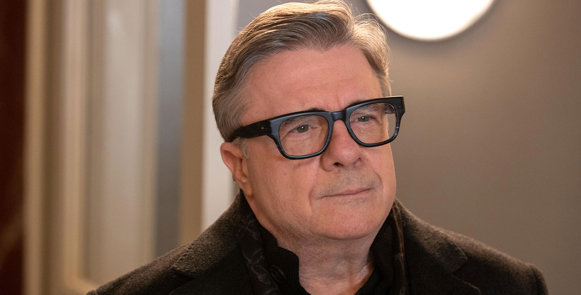 Nathan Lane, in character as Teddy Dimas, wears a wool coat and matching scarf and black glasses. He has pensive expression on his face.