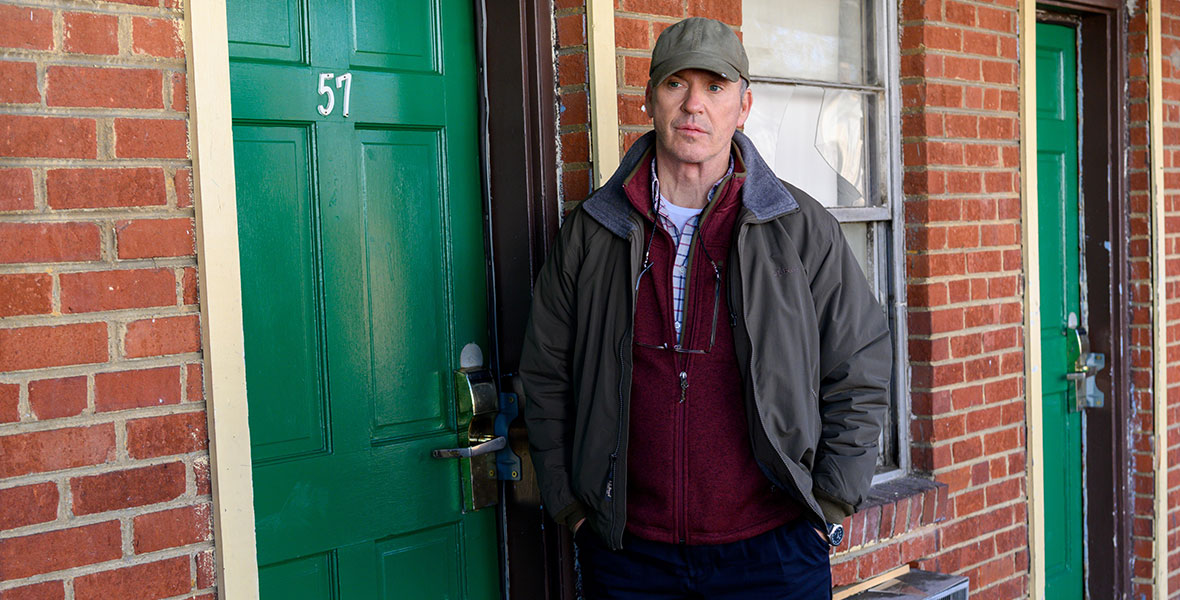 Michael Keaton, in character as Dr. Samuel Finnix, layers a fleece L.L. Bean jacket over a maroon zip-up jacket, a plaid shirt, and a white undershirt. Glasses hang from his neck, and he’s wearing a baseball cap and jeans. He is standing in front of a green door, with the white number 57 on it, and a brick wall.