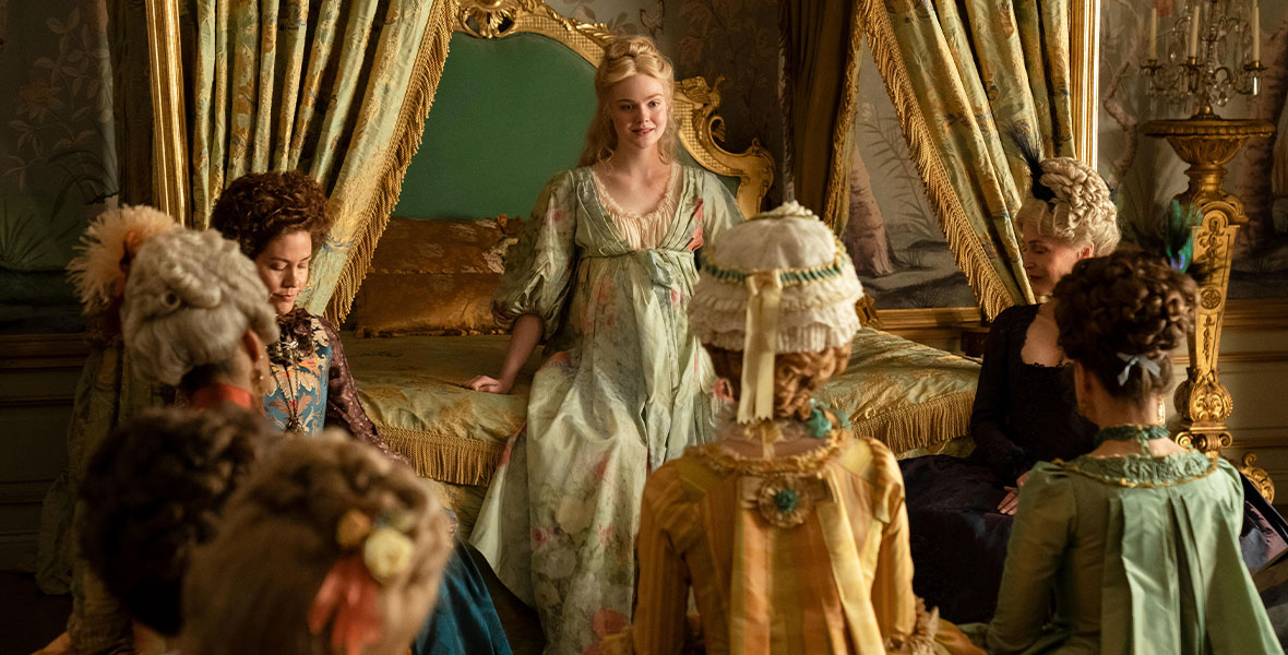 Elle Fanning, in character as Catherine, is seated on the edge of her bed and flanked by Belinda Bromilow, in character as Aunt Elizabeth, and Gillian Anderson, in character as Joanna, and several extras. They are wearing ornate costumes and big wigs.