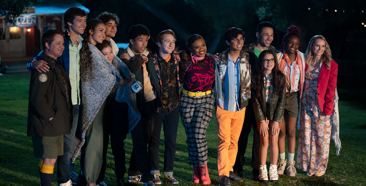 The cast of High School Musical: The Musical: The Series—(L-R) Jason Earles, Matt Cornett, Sofia Wylie, Saylor Bell Curda, Joshua Bassett, Adrian Lyles, Larry Saperstein, Dara Reneé, Frankie Rodriguez, Liamani Segura, Corbin Bleu, Aria Brooks, and Kate Reinders—pose for a group photo outside at Camp Shallow Lake. They have their arms wrapped around each other and are smiling.