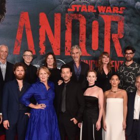 Benjamin Caron, Dan Gilroy, Kyle Soller, Nicholas Britell, Fiona Shaw, Diego Luna, Sanne Wohlenberg, Tony Gilroy, Genevieve O'Reilly, Adria Arjona, and John Gilroy pose on the red carpet at the special three-episode launch event for Lucasfilm's original series Andor at the El Capitan Theatre in Hollywood, California. The group stands in front of a large wall featuring the key art for Andor. On each side of the group are stormtroopers in black and white armor.