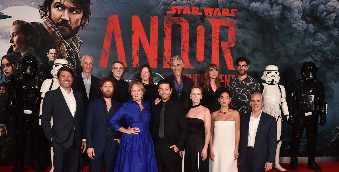 Benjamin Caron, Dan Gilroy, Kyle Soller, Nicholas Britell, Fiona Shaw, Diego Luna, Sanne Wohlenberg, Tony Gilroy, Genevieve O'Reilly, Adria Arjona, and John Gilroy pose on the red carpet at the special three-episode launch event for Lucasfilm's original series Andor at the El Capitan Theatre in Hollywood, California. The group stands in front of a large wall featuring the key art for Andor. On each side of the group are stormtroopers in black and white armor.