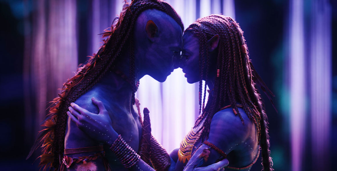 (L-R): The Na’vi avatar Jake Sully (voiced by Sam Worthington) and the Na’vi warrior Neytiri (voiced by Zoe Saldaña) embrace each other and press their foreheads together. Their blue skin is illuminated by the bioluminescent fauna in the background.