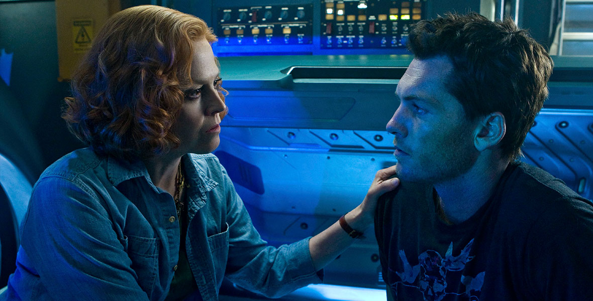 (L-R): Sigourney Weaver, in character as Dr. Grace Augustine, places her left hand on the shoulder of Sam Worthington, in character as Jake Sully. Their skin is illuminated by the blue avatar pod behind them.