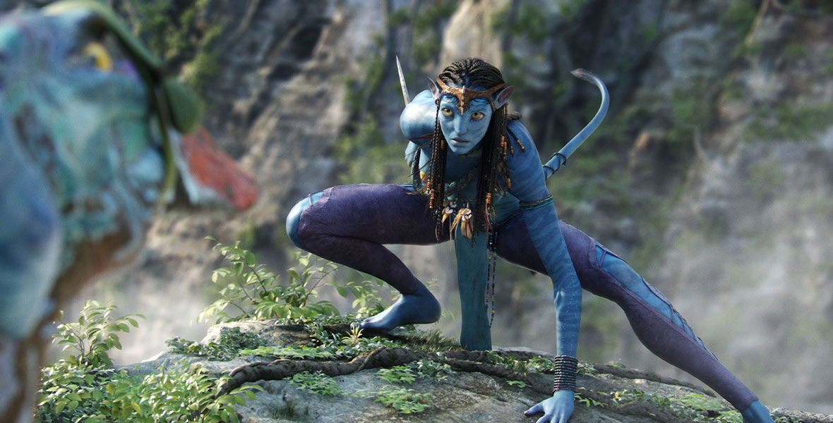 The blue Na’vi warrior Neytiri (voiced by Zoe Saldaña) crouches on a rock. Her tail is extended, and she holds a knife behind her back, ready to strike at any moment.