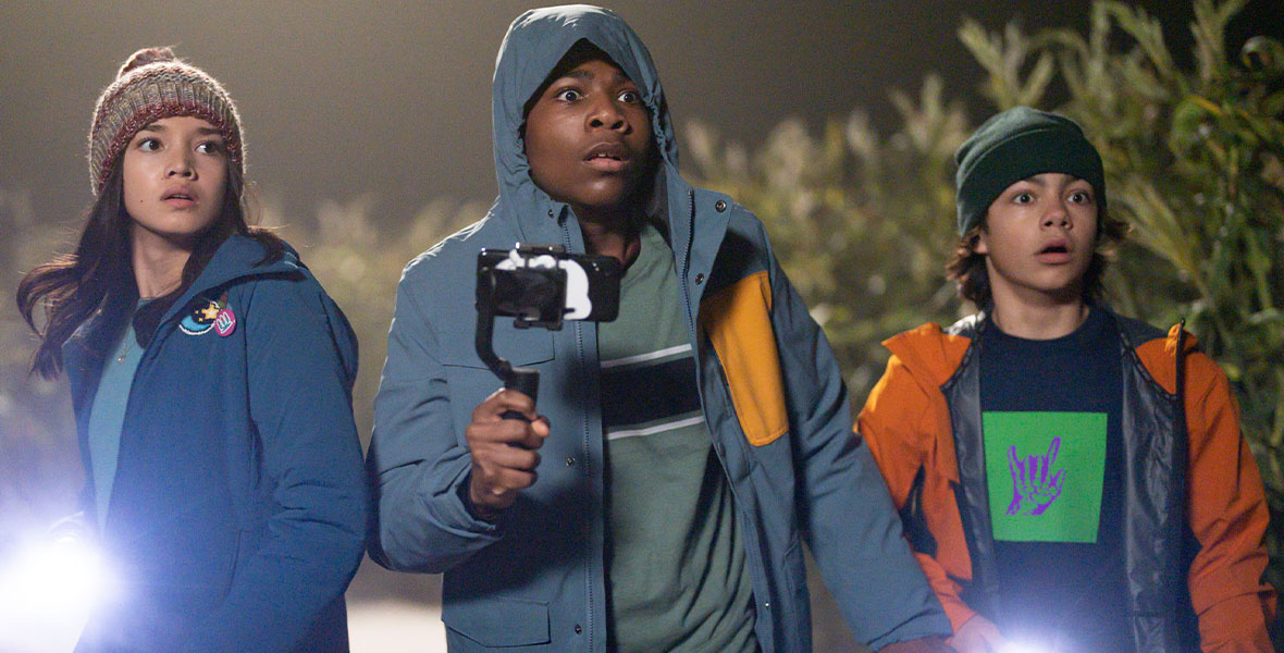 (From left to right): Sophia Hammons, in character as Amy, Christian J. Simon, in character as Gilbert, and Malachi Barton, in character as Marshall, walk through a corn maze at night. They are wearing jackets, hats and pants, and Gilbert is recording a video on his phone. The three of them have scared expressions on their faces.