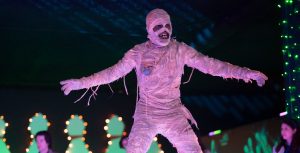 Phil Wright, in character as the mummy Harold, is bandaged and wearing an amulet. He is standing on an illuminated dance floor, with his arms and legs outstretched. In the background, bowling pins are lit up and people are wearing Halloween costumes.