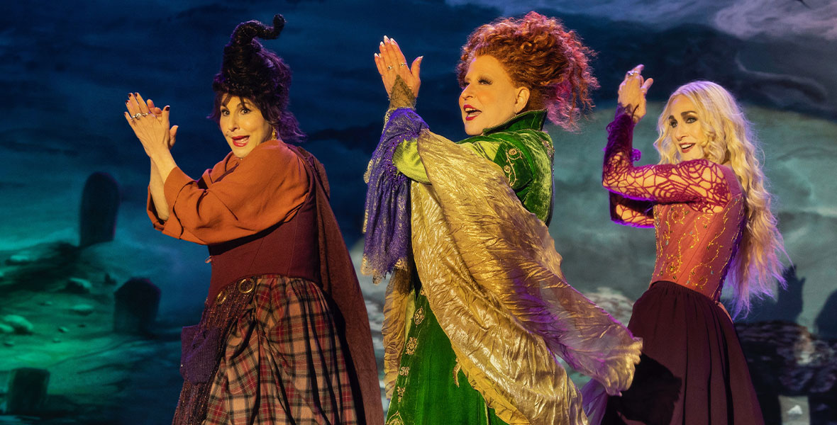From left to right: Kathy Najimy, Bette Midler, and Sarah Jessica Parker in character as Mary Sanderson, Winifred Sanderson, and Sarah Sanderson, respectively. The three witches are onstage and clapping in unison. They are dressed in ornate 17th-century style outfits in their signature colors: red for Mary, green for Winifred, and purple for Sarah.